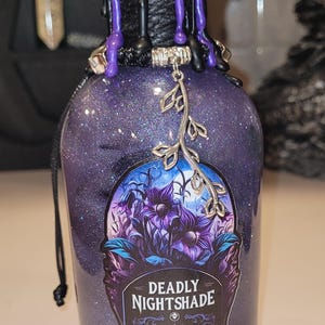 May include: A purple glass bottle with a black wax seal and a silver charm. The bottle is filled with glitter and has a label that reads "Deadly Nightshade".