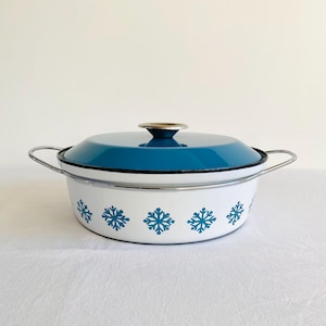 May include: A white enamel casserole dish with a teal lid and snowflake pattern. The dish has silver handles and a gold-coloured knob on the lid. The snowflake pattern is teal. The dish is on a white surface.