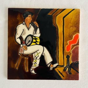 Può includere: Piastrella quadrata in ceramica con un'illustrazione di arte popolare. Un musicista in abito bianco suona uno strumento a fiato e un tamburo. Lo sfondo comprende un camino con tonalità arancioni e marroni e un bordo marrone scuro.