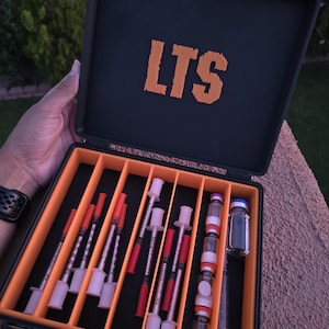 May include: A black carrying case with orange interior dividers and the letters LTS printed in orange on the inside lid. The case contains several medical syringes with red caps and small glass vials with blue and white caps arranged in slots.