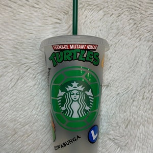 May include: A clear plastic tumbler with a green straw. The tumbler has a Teenage Mutant Ninja Turtles design with the words "Teenage Mutant Ninja Turtles" and "Cowabunga" printed on it. The tumbler also features a green and silver Starbucks logo.