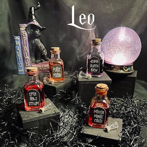 May include: Four small glass bottles with cork stoppers and black labels. The labels have handwritten text that reads "Praise Potion", "Drama Queen Brew", "Spot Light Serum", and "Mirror Magic Elixir". The bottles are arranged on small wooden blocks and surrounded by black, wispy material.
