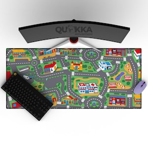 Op de afbeelding: Een kleurrijke muismat met een stadsplattegrond, inclusief wegen, gebouwen en voertuigen. Een zwart toetsenbord en een paarse muis liggen erop. De monitor toont de merknaam "QUKKA MOUSEPADS".