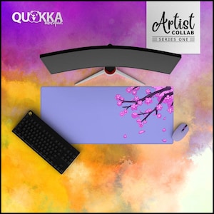 May include: A lavender mouse mat featuring a cherry blossom design and the text "Artist Collab Series One." A black keyboard, a lavender mouse, and a curved monitor are also visible. The mouse mat has the logo "Quokka mousepads."