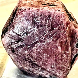 Puede incluir: Una gema rugosa, facetada, de color púrpura rojizo con una estructura cristalina. La gema tiene una forma ligeramente irregular y está colocada sobre un fondo blanco. Una regla es visible en la imagen, proporcionando escala.