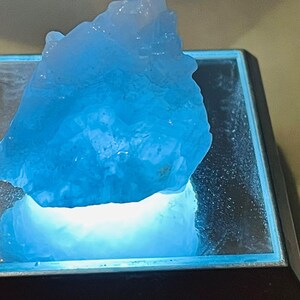 May include: A large, light blue crystal sits on a black square base with a clear glass top. The crystal is illuminated from below, casting a blue glow.