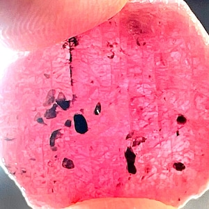 May include: A rough-cut, translucent pink gemstone with dark inclusions. The stone is held between two fingers, showcasing its irregular shape and internal texture. The gem's color ranges from light to deep pink.
