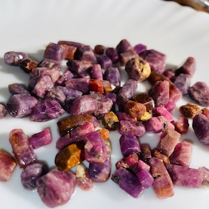 May include: A collection of raw ruby gemstones in various shades of red and purple. The rough-cut stones are scattered on a white surface, showcasing their natural, unpolished textures and crystalline structures. The gemstones vary in size and shape.