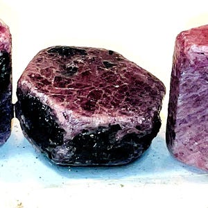 Peut inclure: Trois pierres précieuses brutes, non taillées, roses et violettes avec des inclusions noires. Les pierres précieuses sont facettées et ont un aspect naturel et brut.