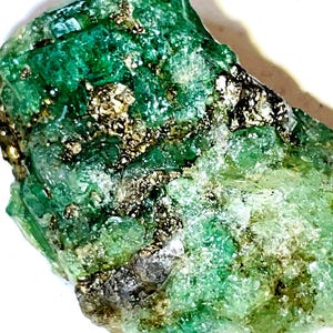 May include: A rough, green emerald gemstone with gold flecks. The emerald is a natural crystal with a rough, irregular shape.
