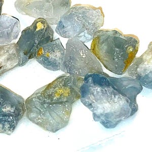 May include: A collection of rough, light blue and gray gemstones with a few flecks of gold. The stones are various shapes and sizes.