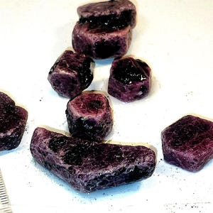 Puede incluir: Un grupo de ocho piedras preciosas brutas de color púrpura. Las piedras son de varias formas y tamaños, algunas tienen una forma hexagonal.