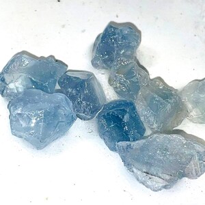 May include: A collection of light blue, translucent, rough-cut aquamarine crystals on a white surface.
