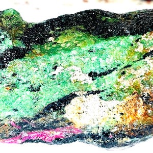May include: A rough, green, black, and brown gemstone with pink accents. The stone has a natural, unpolished look.