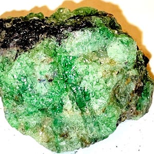 May include: A rough, green and black gemstone with a crystalline texture. The gemstone is about 2 inches in diameter.