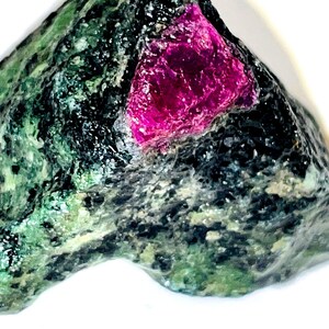 Può includere: Una roccia ruvida di colore verde scuro e nero con un cristallo di rubino rosa brillante incastonato al centro.