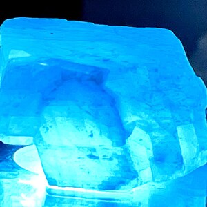 May include: A large, translucent blue crystal with a rough, irregular surface. The crystal is illuminated from below, casting a bright blue glow.