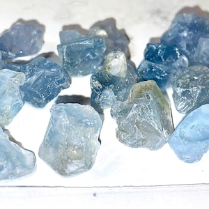 May include: A collection of light blue, translucent, rough-cut gemstones. The stones are various sizes and shapes, with some showing a slight green hue.