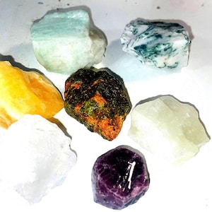 May include: Seven different colored gemstones, including green, yellow, white, orange, and purple, are arranged in a circle on a white surface.