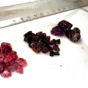 May include: Three piles of pink and red gemstones on a white surface. The gemstones are various sizes and shapes.