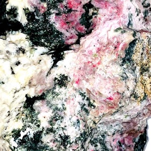 Peut inclure: Gros plan sur un spécimen minéral avec un mélange de couleurs. La roche présente des zones noires profondes, mouchetées de blanc, et des taches roses et rouges. La texture semble rugueuse et inégale.