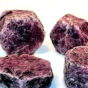 Könnte beinhalten: Vier rohe, violette Rubinkristalle mit einer dunklen, fast schwarzen Farbabweichung. Die Kristalle sind facettiert und haben ein natürliches, unpoliertes Aussehen.