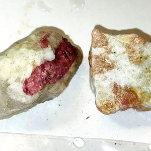 Peut inclure: Deux spécimens minéraux bruts, blancs et roses, avec des inclusions rouges et orange. Les spécimens sont sur une surface blanche.