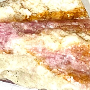 Peut inclure: Un cristal brut rose et blanc naturel avec des stries brunes et orange. Le cristal a un aspect stratifié.