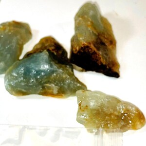 May include: A group of four rough, green and brown gemstone pieces. The stones are translucent and have a slightly cloudy appearance.