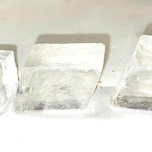 May include: Three clear, translucent, cubic crystals. The crystals are smooth and have a slightly cloudy appearance.