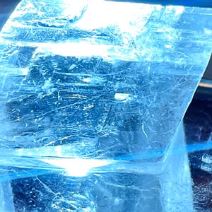 May include: A large, clear, crystalline rock with a light blue hue. The rock is sitting on a black display stand.