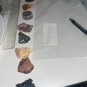 Puede incluir: Una colección de nueve piedras de diferentes colores, incluyendo una amarilla, una morada, una rosa, una verde, una naranja claro, una roja, una negra, una gris oscuro y una gris claro. Las piedras están dispuestas en una fila sobre una superficie blanca. El texto "SKJ4RCEOKS" está escrito en un papel blanco.