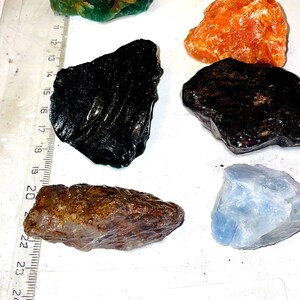 Puede incluir: Colección de piedras preciosas en bruto de varios colores: verde, naranja, negro, marrón y azul. Las piedras tienen formas irregulares y parecen naturales. Una regla graduada en centímetros está al lado. El texto "CGQSCPC" está escrito.