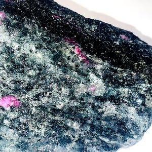 May include: A dark gray rock with pink ruby crystals embedded in it. The rock has a rough, uneven surface.