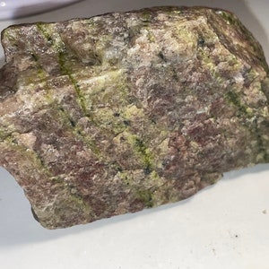 May include: A rough, irregularly shaped rock with a mix of green, brown, and pink hues. The rock has a layered appearance with streaks of green running through it.
