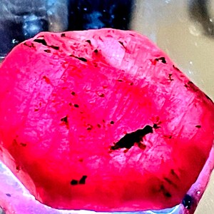May include: A large, deep red ruby gemstone with a rough, faceted surface. The ruby has black inclusions throughout.