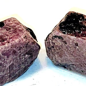 Peut inclure: Deux cristaux de rubis bruts, rose foncé et noirs. Les cristaux sont de forme irrégulière et ont une surface rugueuse et texturée.