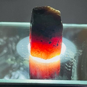 Puede incluir: Una piedra preciosa de color rojo oscuro y negro con un acabado rugoso y natural. La piedra preciosa está iluminada desde abajo, destacando su color rojo intenso.