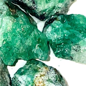 May include: A collection of raw emerald crystals, showcasing their natural green color and rough, unpolished texture. The crystals are various sizes and shapes, with some displaying a slight shimmer.