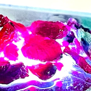 Może przedstawiać: Gromada żywych różowych i fioletowych kamieni szlachetnych, przypominająca stos surowych kryształów, na rozmytym tle turkusowym i białym.