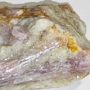 Peut inclure: Un cristal brut, naturel, rose et blanc avec des inclusions jaunes et brunes. Le cristal a une texture stratifiée et striée.