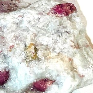 Peut inclure: Une roche blanche avec de petits cristaux rouges incrustés. La roche mesure environ 5 cm de large et 7 cm de long.
