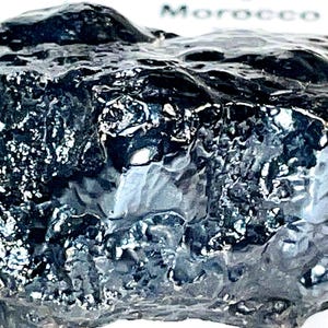 Pode incluir: Uma rocha escura, de forma irregular, com uma superfície brilhante e texturizada. A rocha tem uma cor preta profunda com reflexos. A palavra "Morocco" é visível no fundo. Este item é provavelmente um espécime mineral ou uma pedra decorativa.