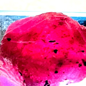 May include: A large, deep red ruby gemstone with black inclusions. The ruby is rough and unpolished, with a natural, irregular shape.