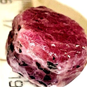 May include: A large, rough, pink ruby gemstone with black inclusions. The ruby is irregularly shaped and has a shiny, polished surface.