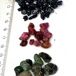 Può includere: Tre gruppi di pietre preziose: nere, rosa e verdi. Le pietre preziose sono disposte in linea verticale su una superficie bianca con un righello sullo sfondo.