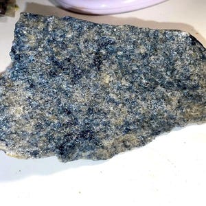 Könnte beinhalten: Nahaufnahme eines rauen, grauen und blauen Steins mit einer geschichteten Textur. Der Stein hat ein gesprenkeltes Aussehen mit kleinen, dunkelblauen Flecken.