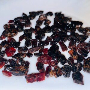 Puede incluir: Una colección de pequeñas piedras preciosas ásperas de color rojo y negro. Las piedras preciosas tienen una forma irregular y un aspecto natural sin pulir.
