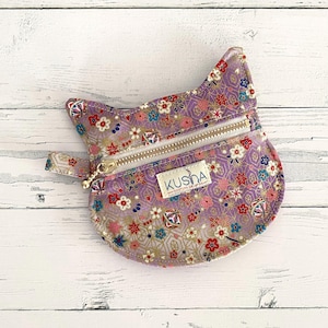 May include: A cat-shaped coin purse in a light purple fabric featuring a repeating pattern of small colourful flowers and geometric shapes. The pouch includes a gold-toned metal zipper closure and a small white fabric label with the text KUSHA.