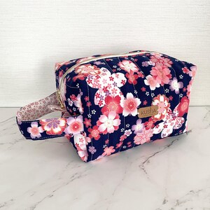 以下が含まれることがあります： 濃い青色の背景にピンクと白の桜の花柄が鮮やかなキルティング生地のポーチ。サイドハンドル、ゴールドカラーのファスナー、KUSHAと刻印された小さな長方形の茶色のレザータグが付いています。